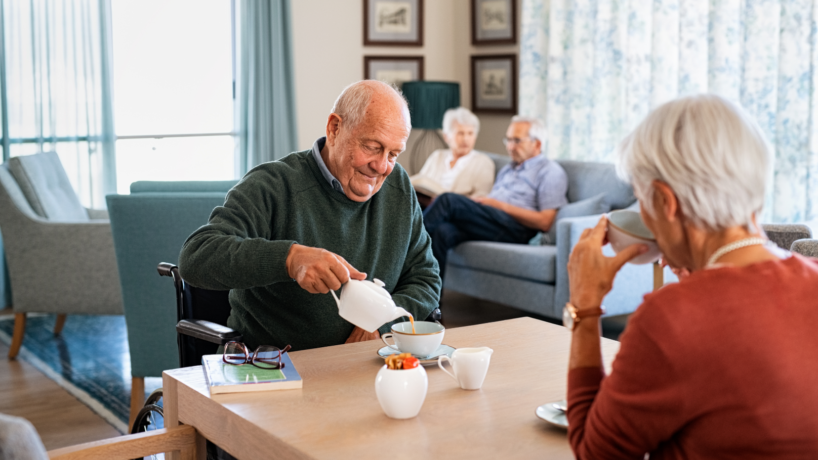 Photo of an elderly couple seated at a table: a man pouring himself a cup of tea while a woman drinks from a cup. In the background is a different elderly couple seated on a couch.