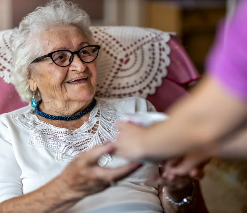 Photo of an elderly woman who is being handed a cup of tea by another person. Only the arms of the other person are visible.