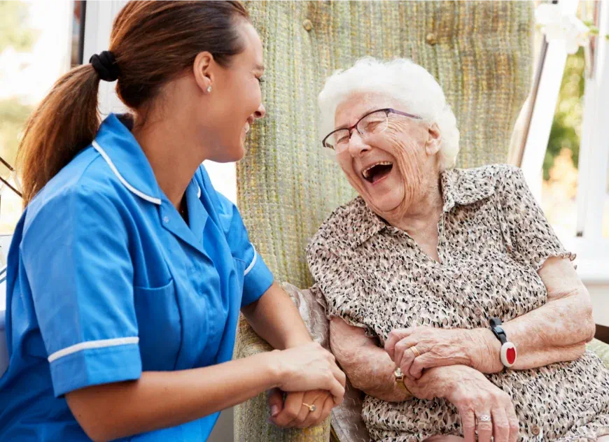 Photo of an elderly woman in an armchair laughing with a nurse who is crouched by her side