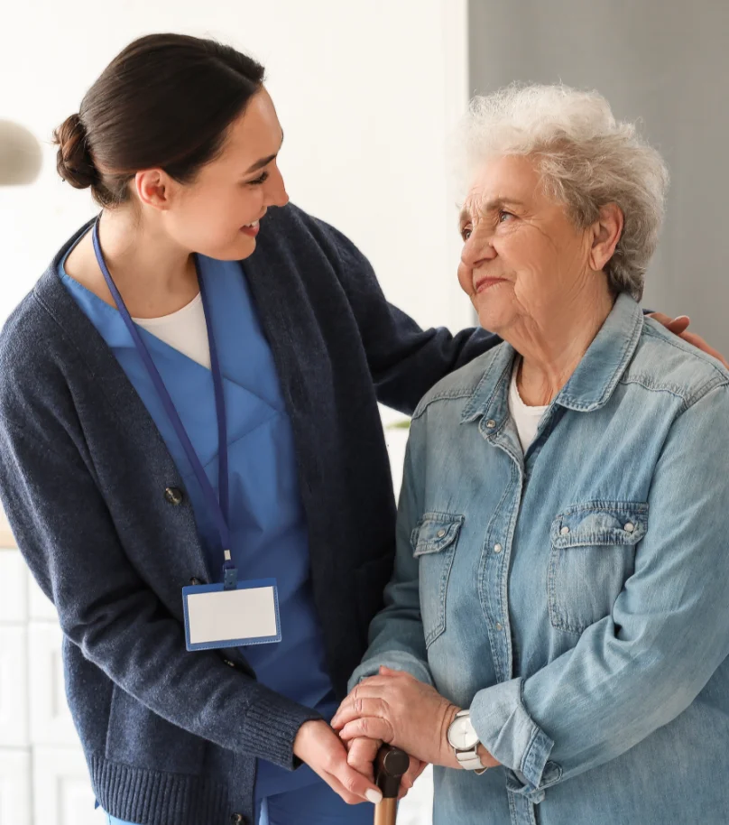 Photo of an elderly woman in a denim shirt looking at a smiling nurse whilst holding a cane
