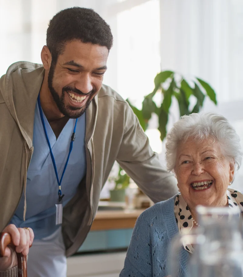 Photo of a smiling male nurse stood next to a smiling, seated elderly woman