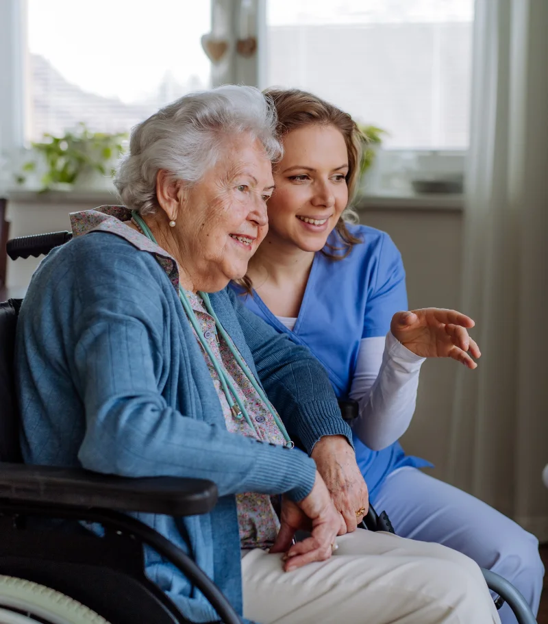 Photo of an elderly woman in a wheelchair, with a nurse crouching next to her. The nurse appears to be pointing at an object that both are looking at.