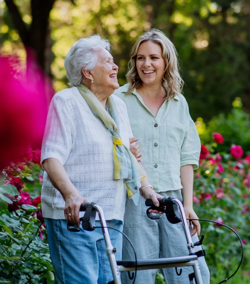 Photo of an elderly woman using a walker, stood next to a younger woman who is holding her arm, both of whom appear to be walking through a garden