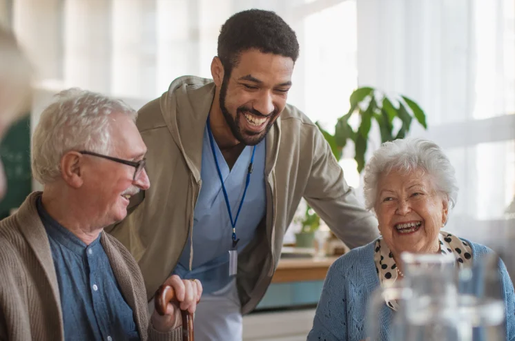 Photo of a male nurse stood next to a seated elderly couple, all of whom are smiling