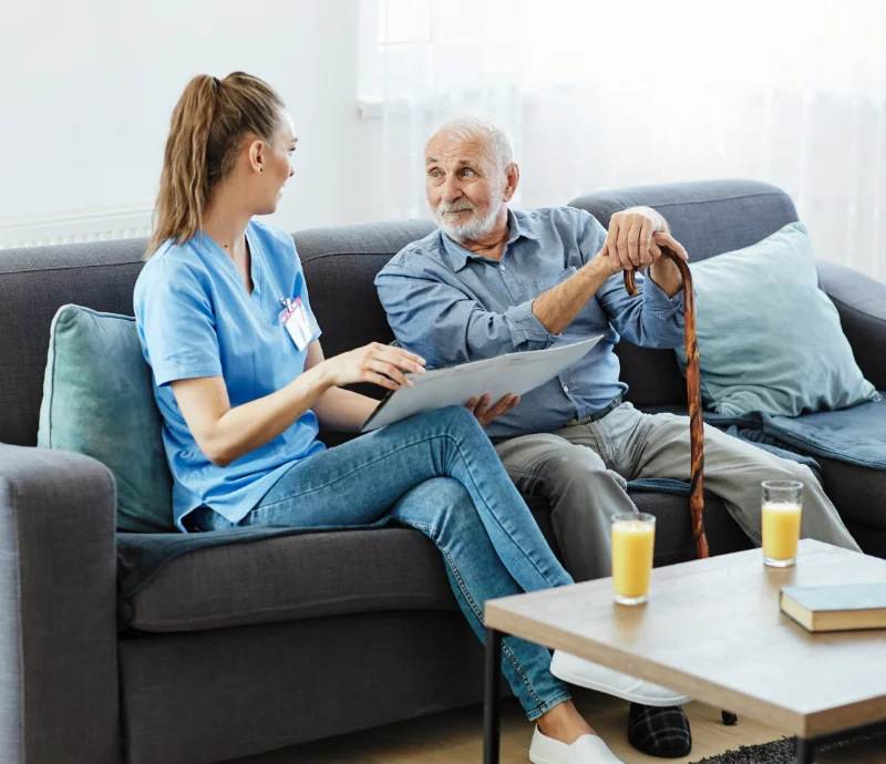 Photo of a man holding a cane looking at a nurse who is holding an open book and appears to be speaking. In front of them is a table upon which are sat two glasses of orange juice and a book.