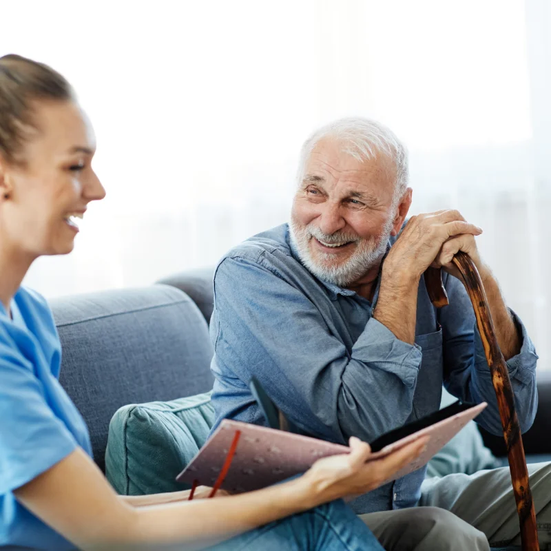 Photo of a man with a cane smiling with a nurse who is holding an open book with a pink cover, with both seated on a couch