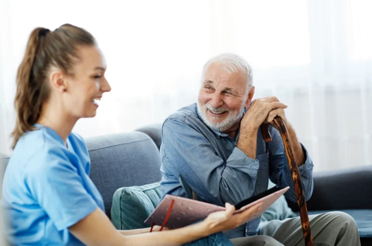 Photo of a man with a cane smiling with a nurse who is holding an open book with a pink cover, with both seated on a couch