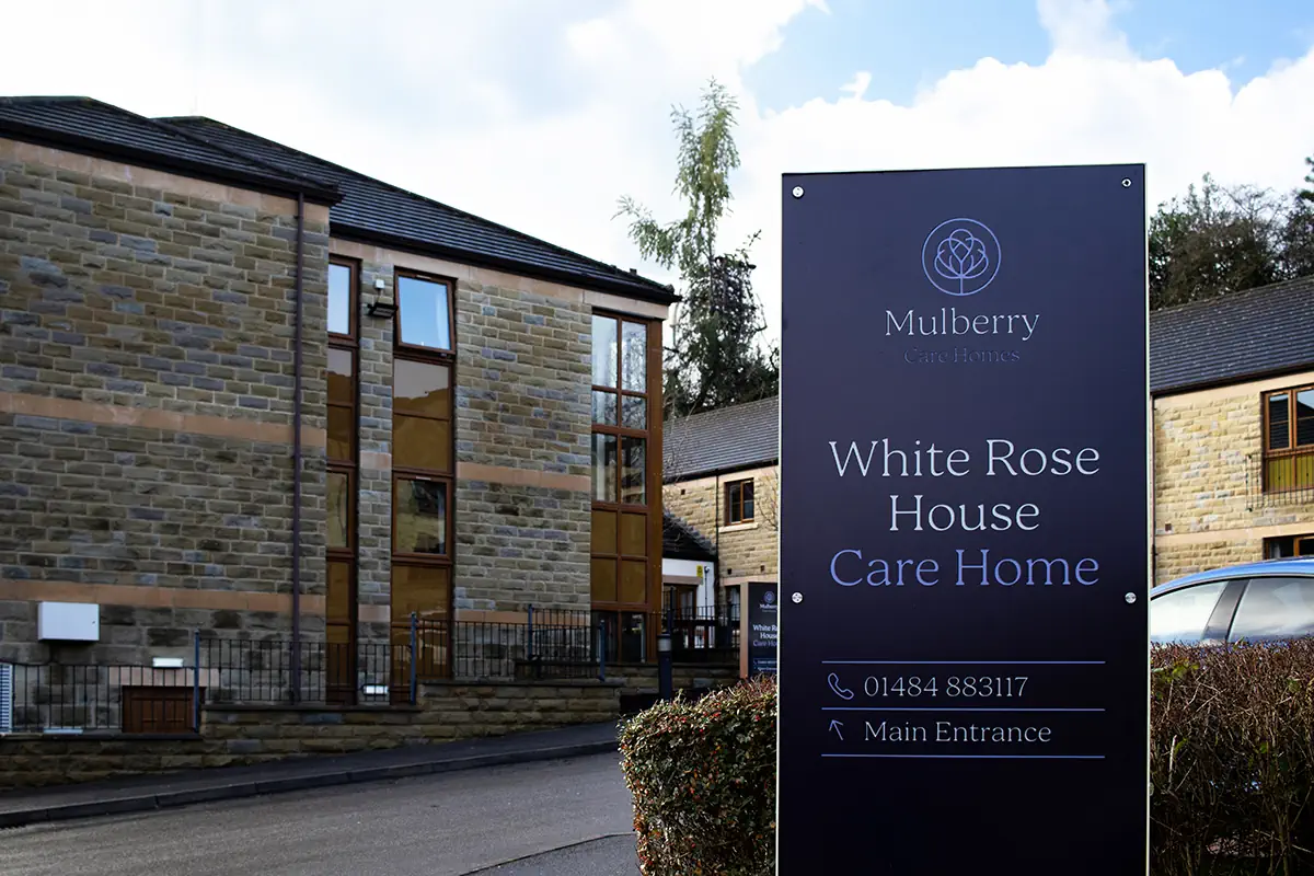 Photo of a sign outside White Rose House Care Home, taken from Huddersfield Road, with the care home buildings visible in the background