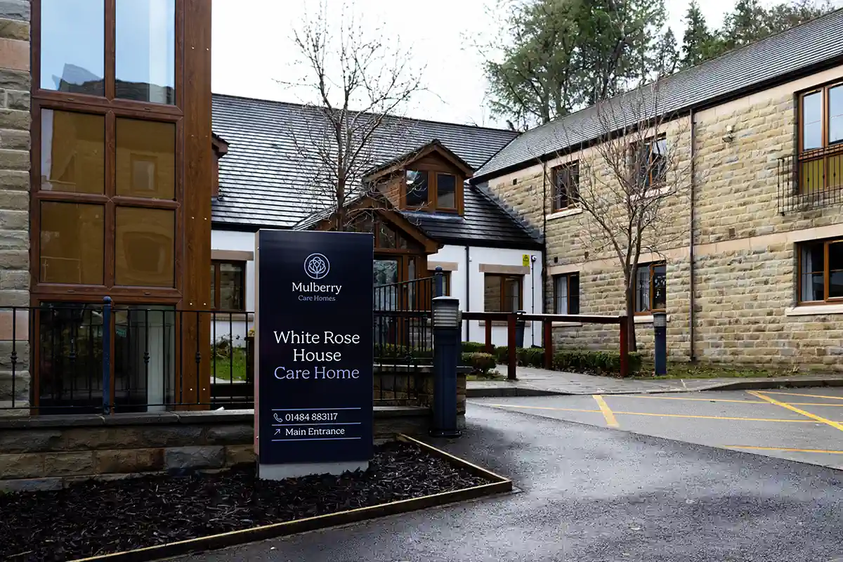 Photo of a sign outside the main entrance of White Rose House Care Home, with the care home buildings visible in the background