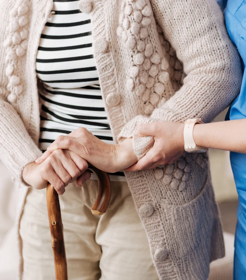 Photo of a woman with a cane being supported by a nurse, neither of whose faces are visible