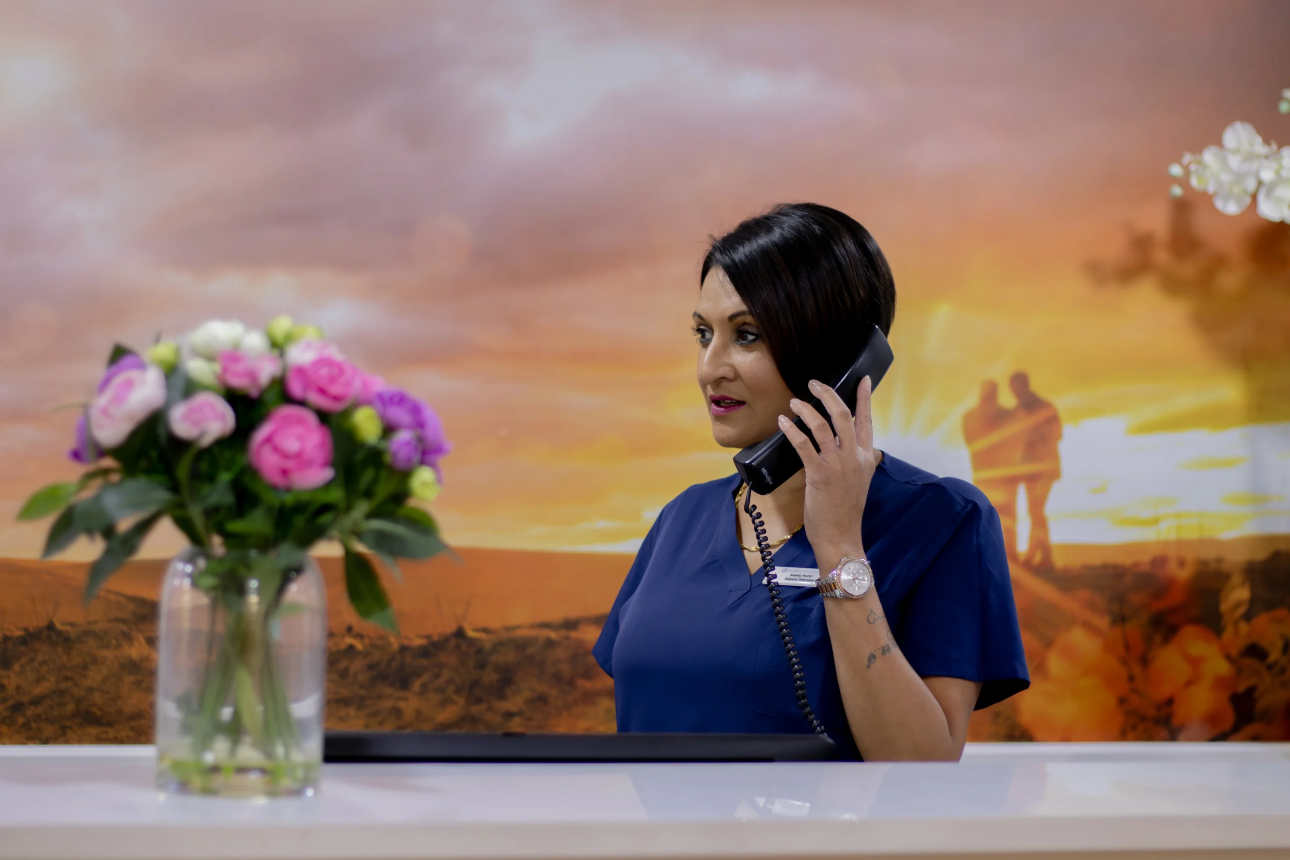 A receptionist standing at a desk in a care home, speaking on the phone with a vase of flowers beside her