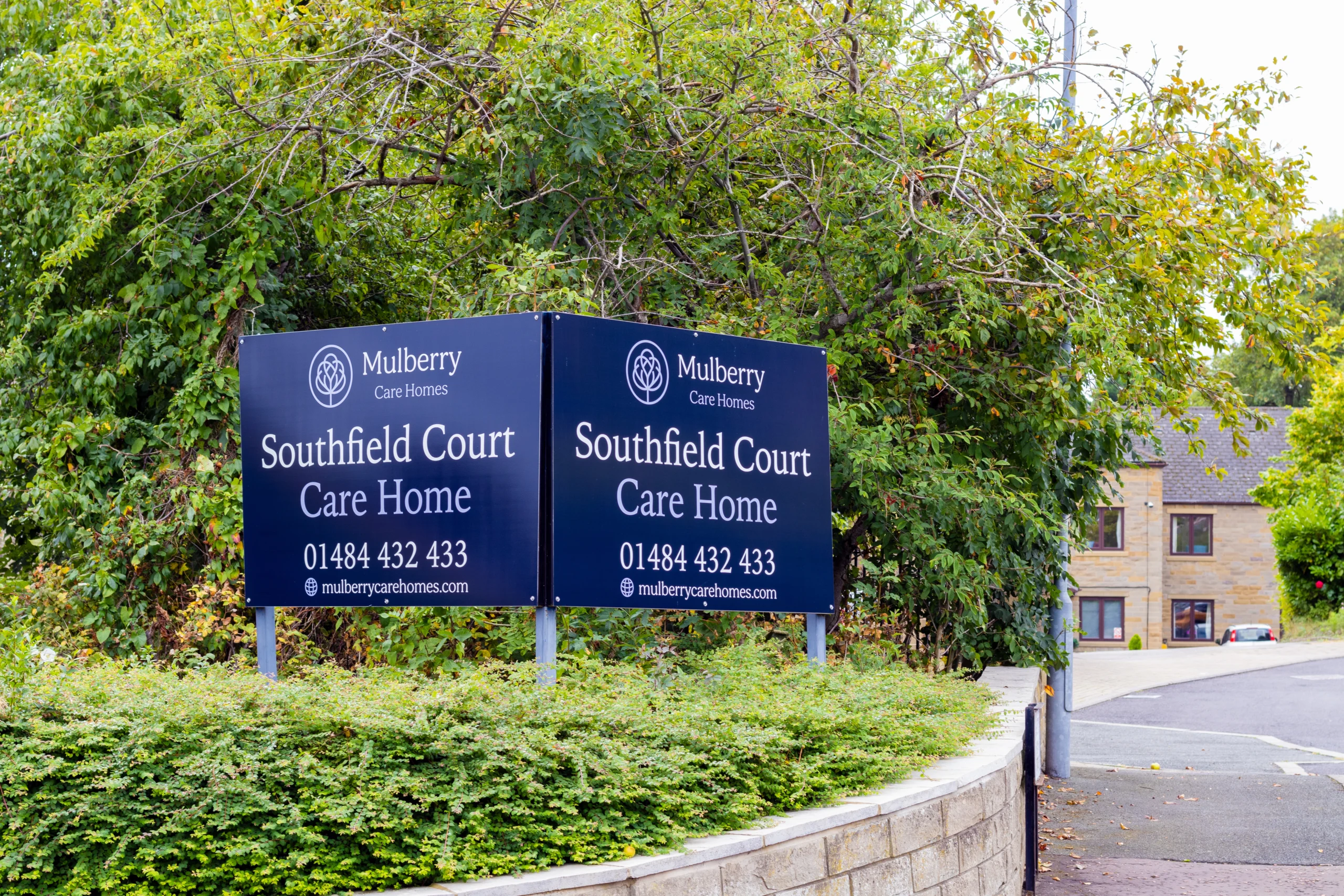 A blue sign for Southfield Court Care Homes, in front a large green bush. In the background is the building for the care home.